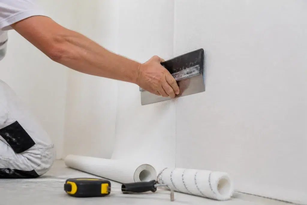 vakman die een baan renovlies met een spatel strak tegen een hoge lichte muur drukt in een nieuwbouwwoning